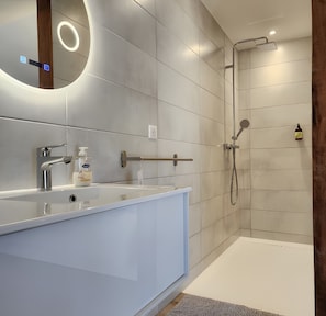 Un baño moderno con tocador blanco, espejo, cabezal de ducha manual y dispensador de jabón.