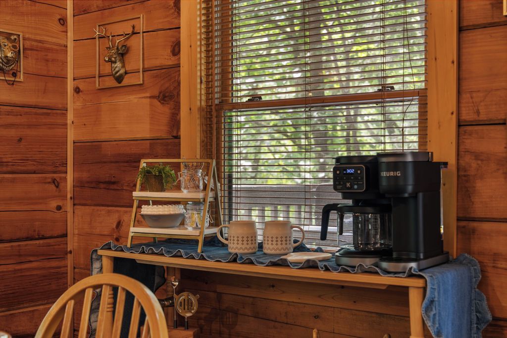 A wooden cabin interior with a coffee station, a shelf with decorative items, and a window with blinds.