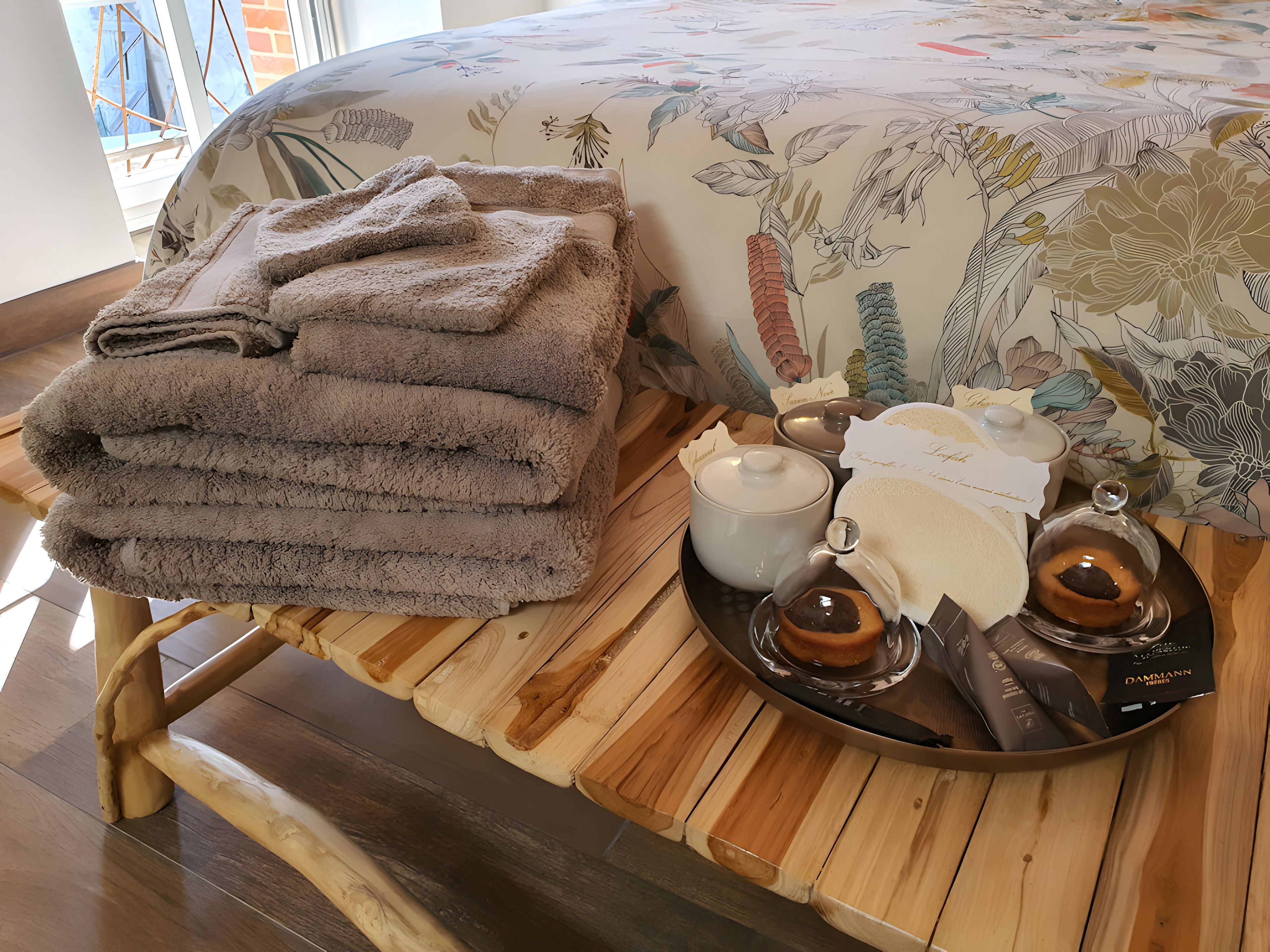 Une pile de serviettes soigneusement pliées, posée sur un tabouret en bois, un plateau avec une théière, des tasses et un récipient en verre, et un lit recouvert d’une couvre-lit à motifs floraux.