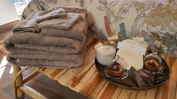 Une pile de serviettes soigneusement pliées, posée sur un tabouret en bois, un plateau avec une théière, des tasses et un récipient en verre, et un lit recouvert d’une couvre-lit à motifs floraux.