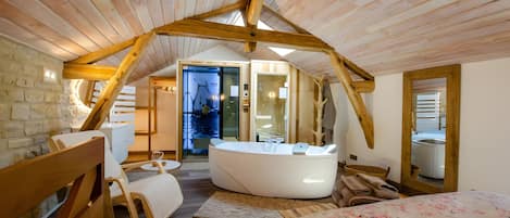 A cozy bedroom with a sloped ceiling, a large bed, a sitting area with a chair and a table, and a bathroom with a bathtub.