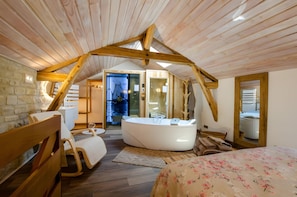 A cozy bedroom with a sloped ceiling, a large bed, a sitting area with a chair and a table, and a bathroom with a bathtub.