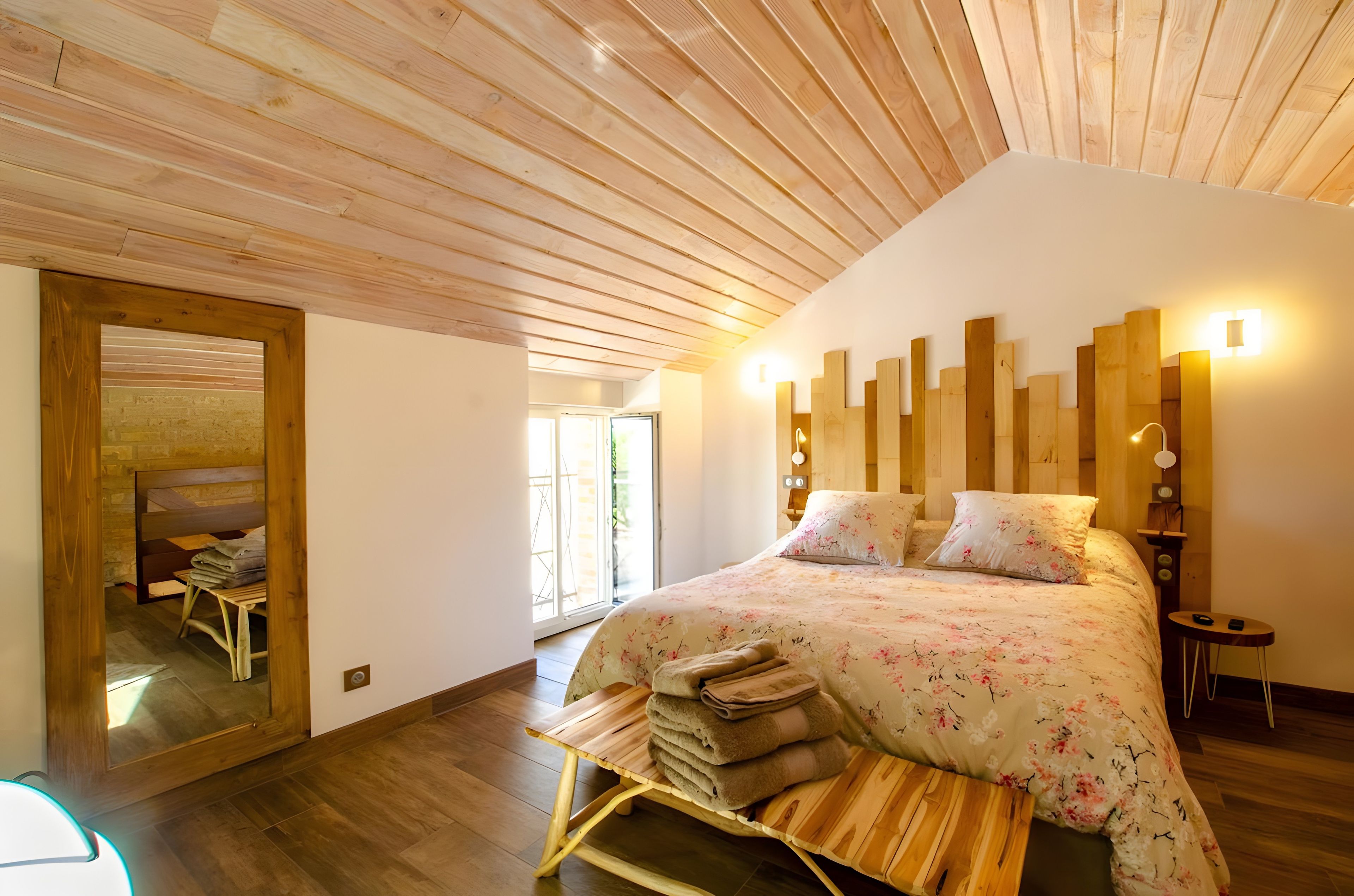 Une chambre avec un plafond en bois, un lit recouvert d’une literie à motifs floraux, une table de chevet en bois et un miroir.