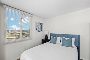 A bedroom with a bed, a nightstand, a lamp, a painting, and a window with blinds. - Azure Horizon - 3 BR Ocean Views & Resort Pool (Marina del Rey)