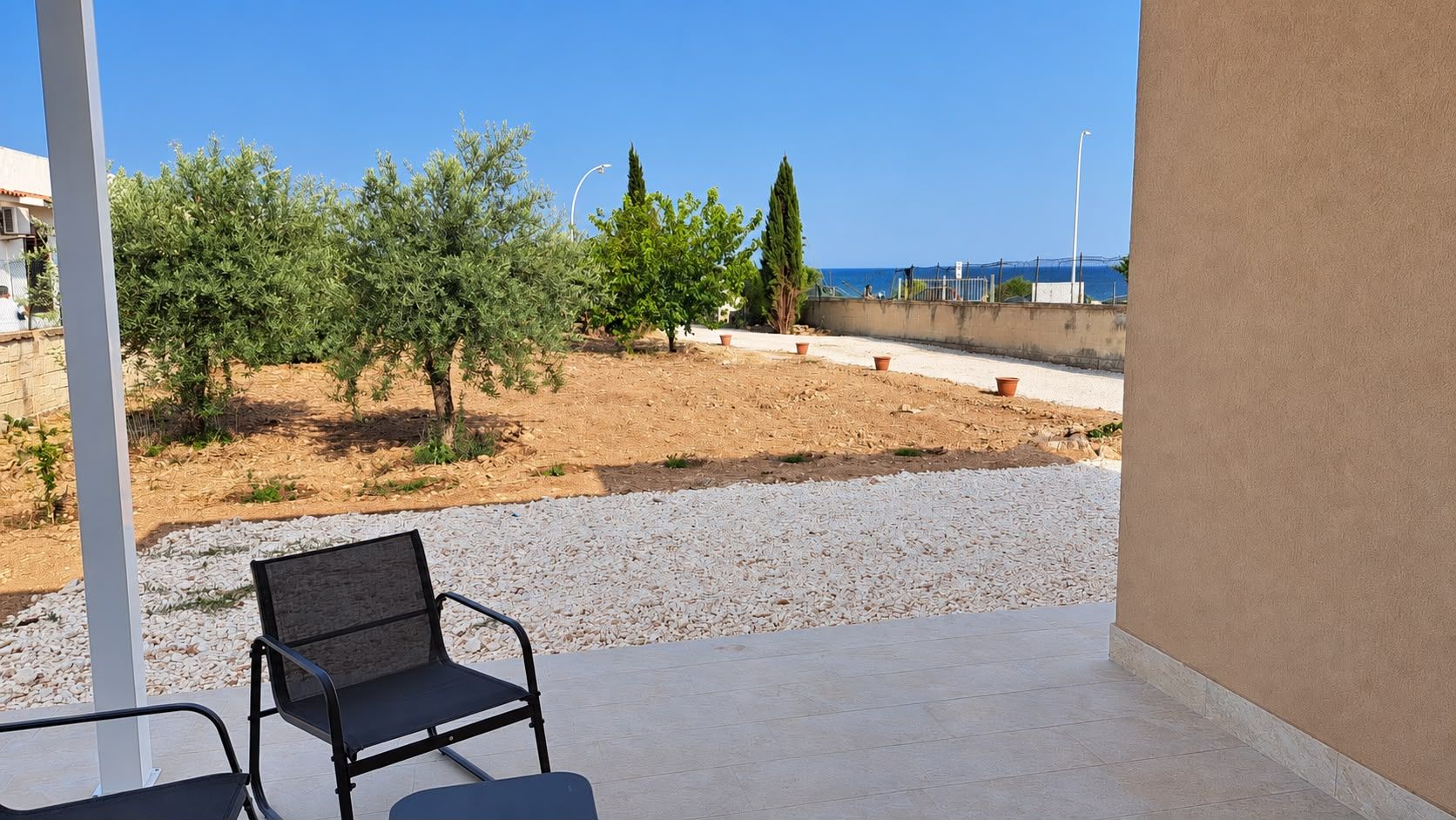 A balcony with a chair, a view of a sandy area with trees, and a clear sky.