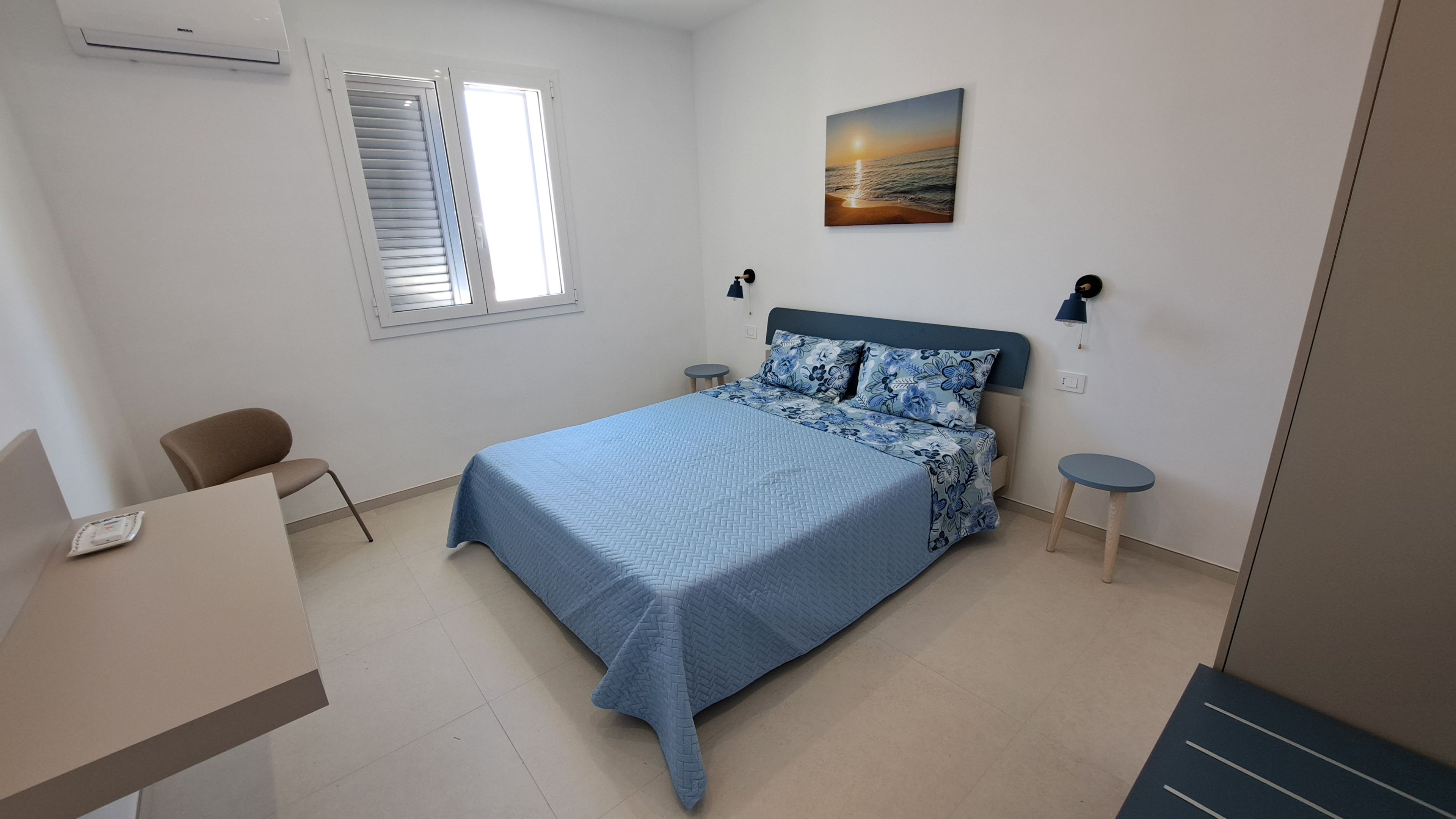 A bedroom with a bed, a chair, a small table, a window with blinds, and a wall-mounted lamp.