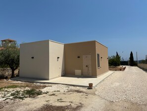 Un edificio moderno de dos pisos con techo plano, una pequeña entrada con puerta y un espacio exterior visible con grava y algunas plantas en macetas.