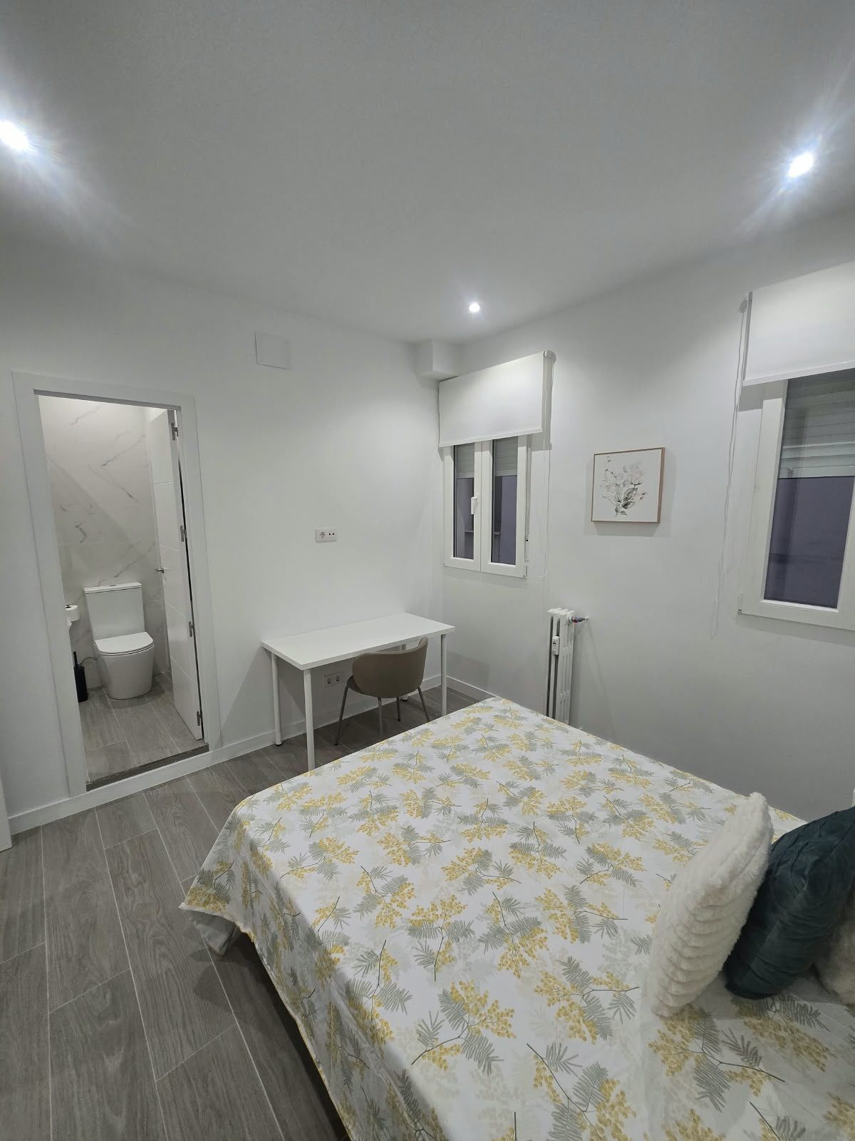 A bedroom with a bed, a desk, a chair, a toilet, and a window.