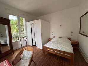 A bedroom with a wooden bed, a wicker chair, a wardrobe, and a window with a view of greenery. - Large family villa 50m from the beach (Arcachon)