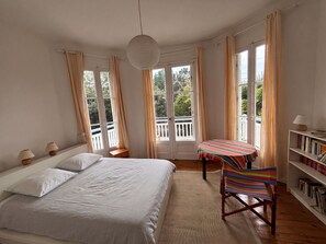 A bedroom with a bed, a small table with a striped tablecloth, a chair, a bookshelf, and a view of the outdoors through large windows. - Large family villa 50m from the beach (Arcachon)