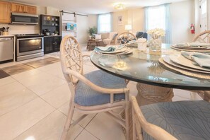 A modern kitchen with built-in appliances, a dining area with a glass table, wicker chairs, and a cozy living space with a sofa and a TV. - West End PCB | 1/2 Block to Beach+Heated Pool (Panama City Beach)