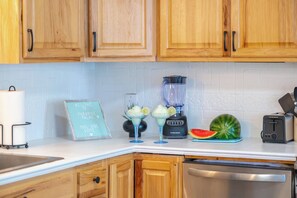A kitchen with wooden cabinets, a blender, a watermelon, a toaster, and a sign that reads 'Welcome to Paradise Palms Pool Side'. - West End PCB | 1/2 Block to Beach+Heated Pool (Panama City Beach)
