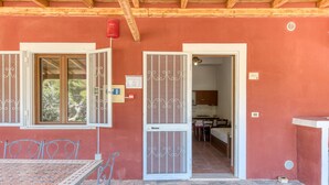 A room with a stone floor, a table with chairs, a window with a view of trees, and a door leading to a kitchen. - Apartment 'Le Colonie - Bilocale 2' with Private Terrace, Shared Garden and Air Conditioning (Presicce)
