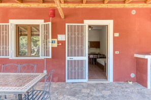 Una stanza con pavimento in pietra, un tavolo con sedie, una finestra vista alberi e una porta che dà su una cucina.