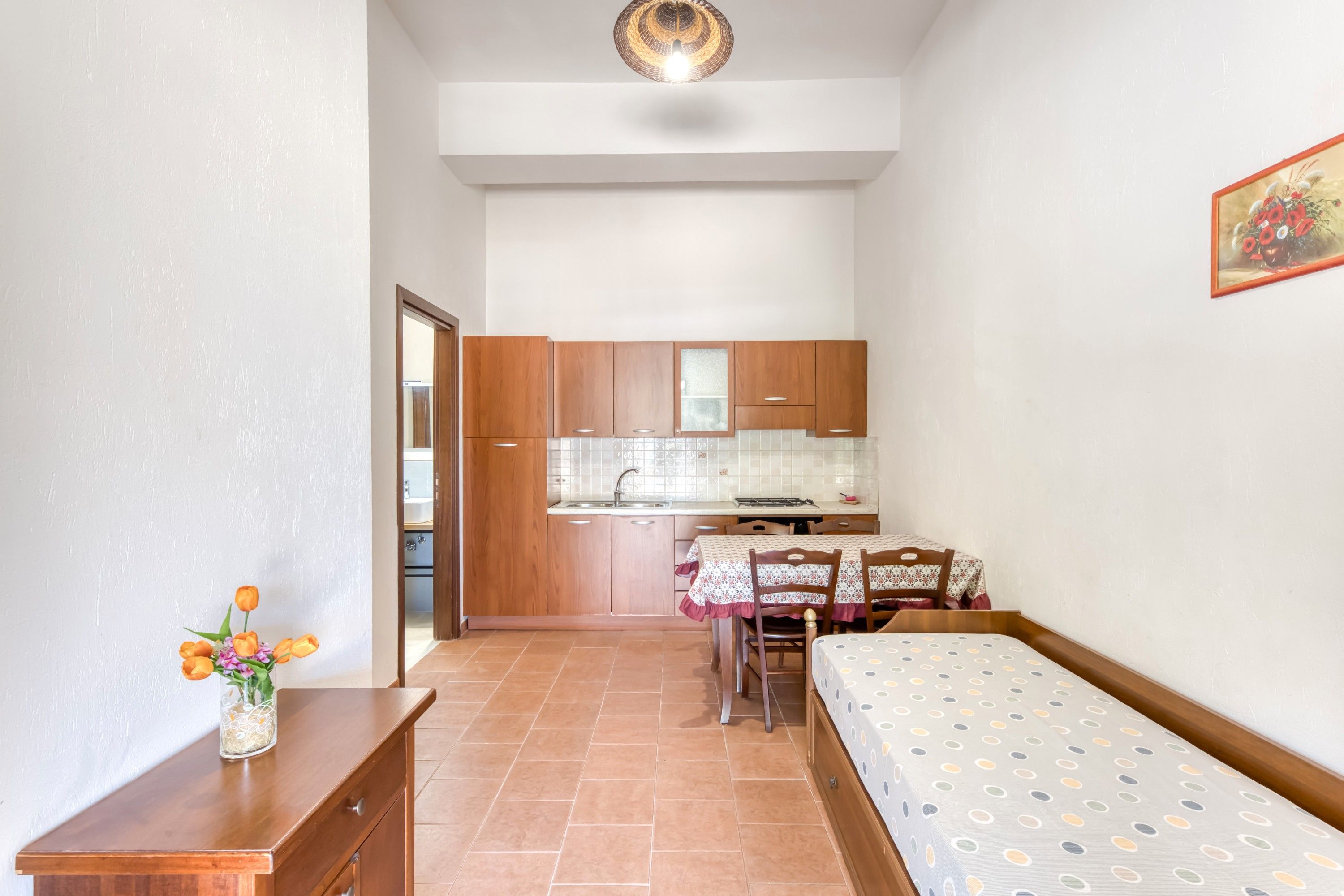 Una stanza compatta con un letto, un tavolino con un vaso di fiori, un tavolo da pranzo con sedie e un angolo cottura con pensili e un lavello.