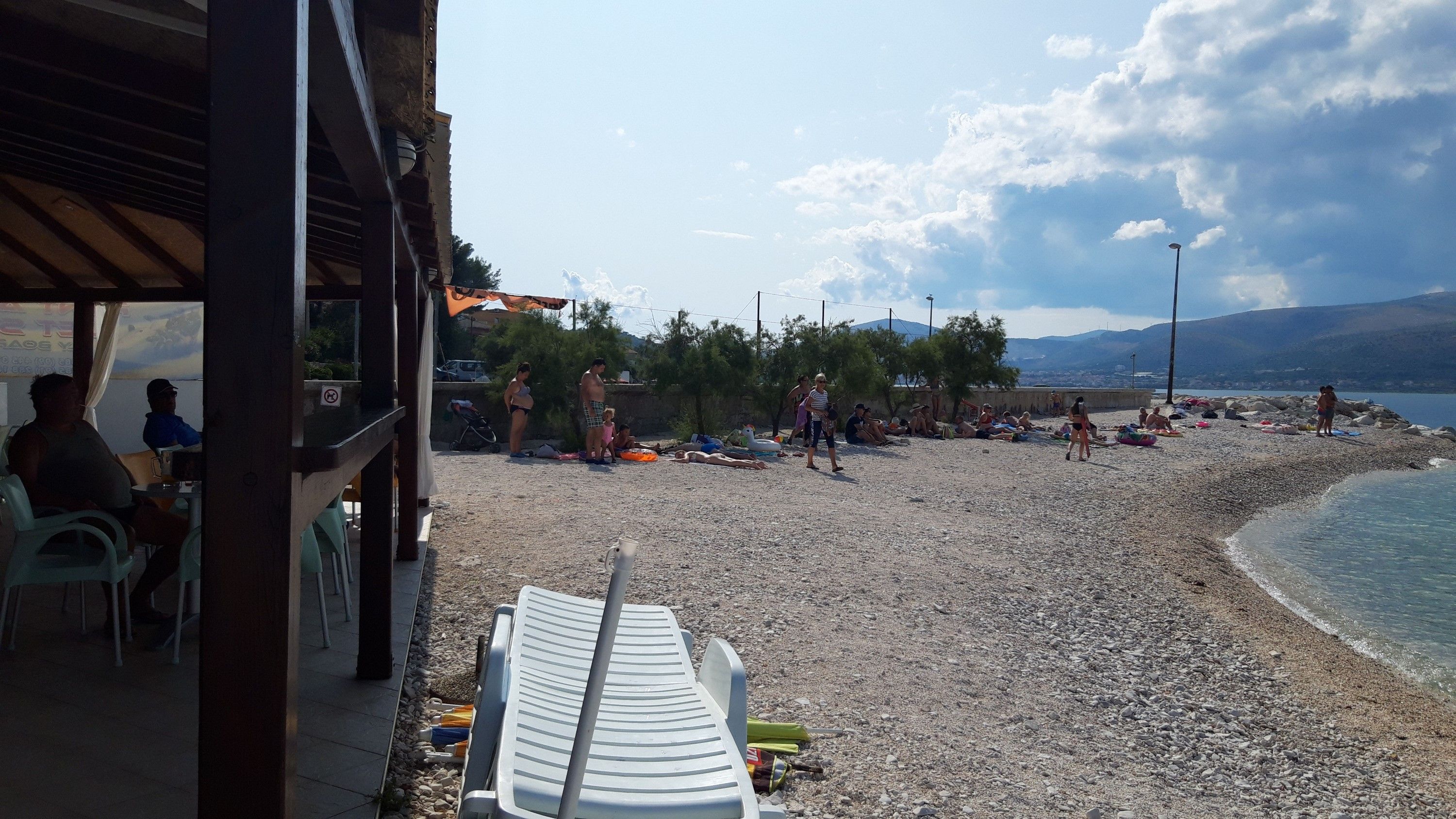 A beach with people sunbathing and swimming, a covered seating area, and a view of the sea and mountains in the background.
