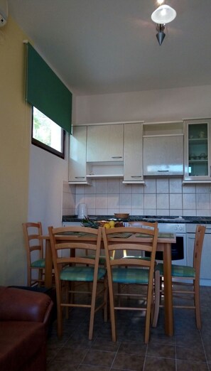 Een kleine keuken met witte kastjes, een eettafel met stoelen, een koffiezetapparaat en een raam met een groene zonwering.