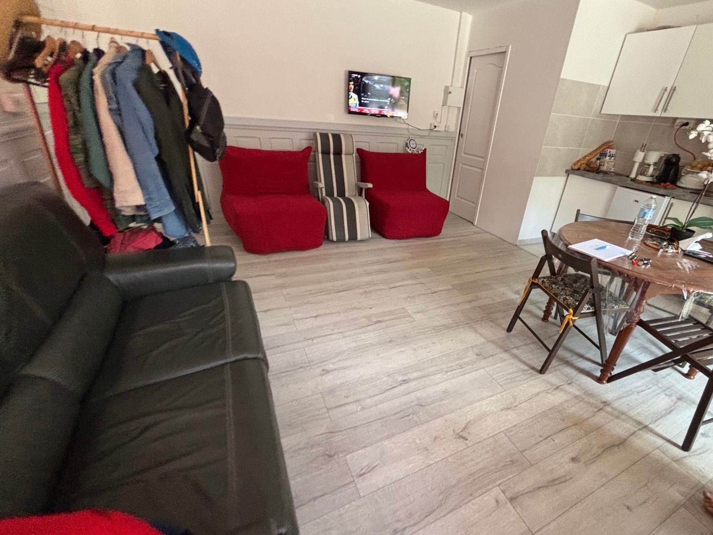 A living room with a black sofa, red chairs, a wooden dining table with chairs, a TV mounted on the wall, and a coat rack with various jackets.