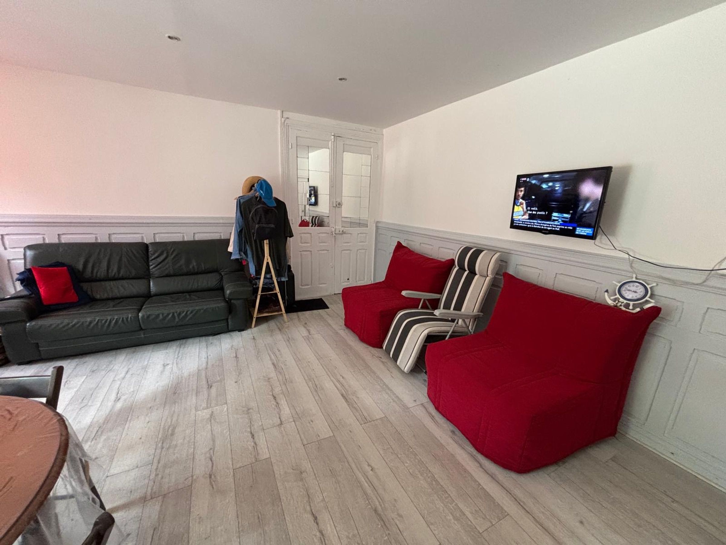 Una sala de estar con un sofá, un sillón rojo, un televisor montado en la pared y piso de madera.