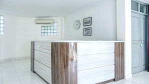 Un mostrador de recepción con un reloj y cuadros en la pared, una ventana con persianas, y una puerta con cerradura.