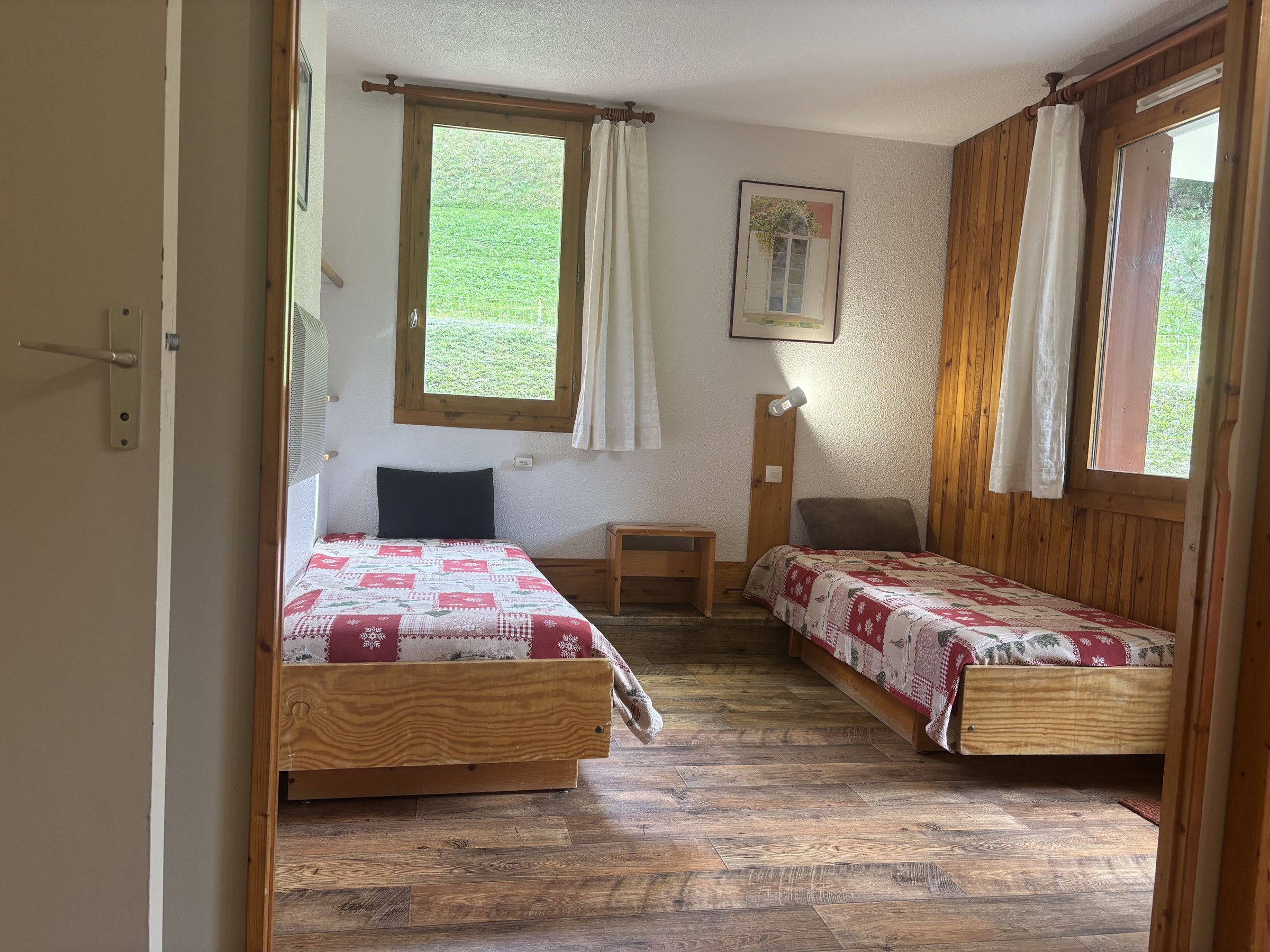 Una habitación con dos camas de madera, un pequeño banco de madera, una mesita de noche y una ventana con cortinas.