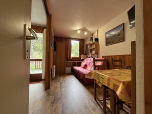 Ein gemütliches Zimmer mit Holzboden, einem Bett mit rosa Decke, einem Tisch mit Tischdecke, einem Bücherregal und einem Fenster mit Vorhängen.