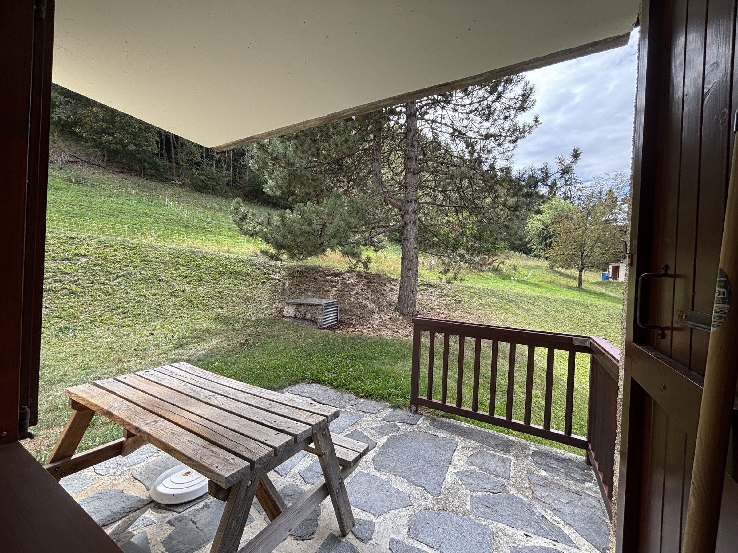 A wooden table on a stone patio with a view of a grassy hill and trees.