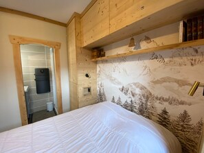 Een slaapkamer met een bed, houten hoofdeinde, een plank met boeken, en een zichtbare badkamer door een open deur.
