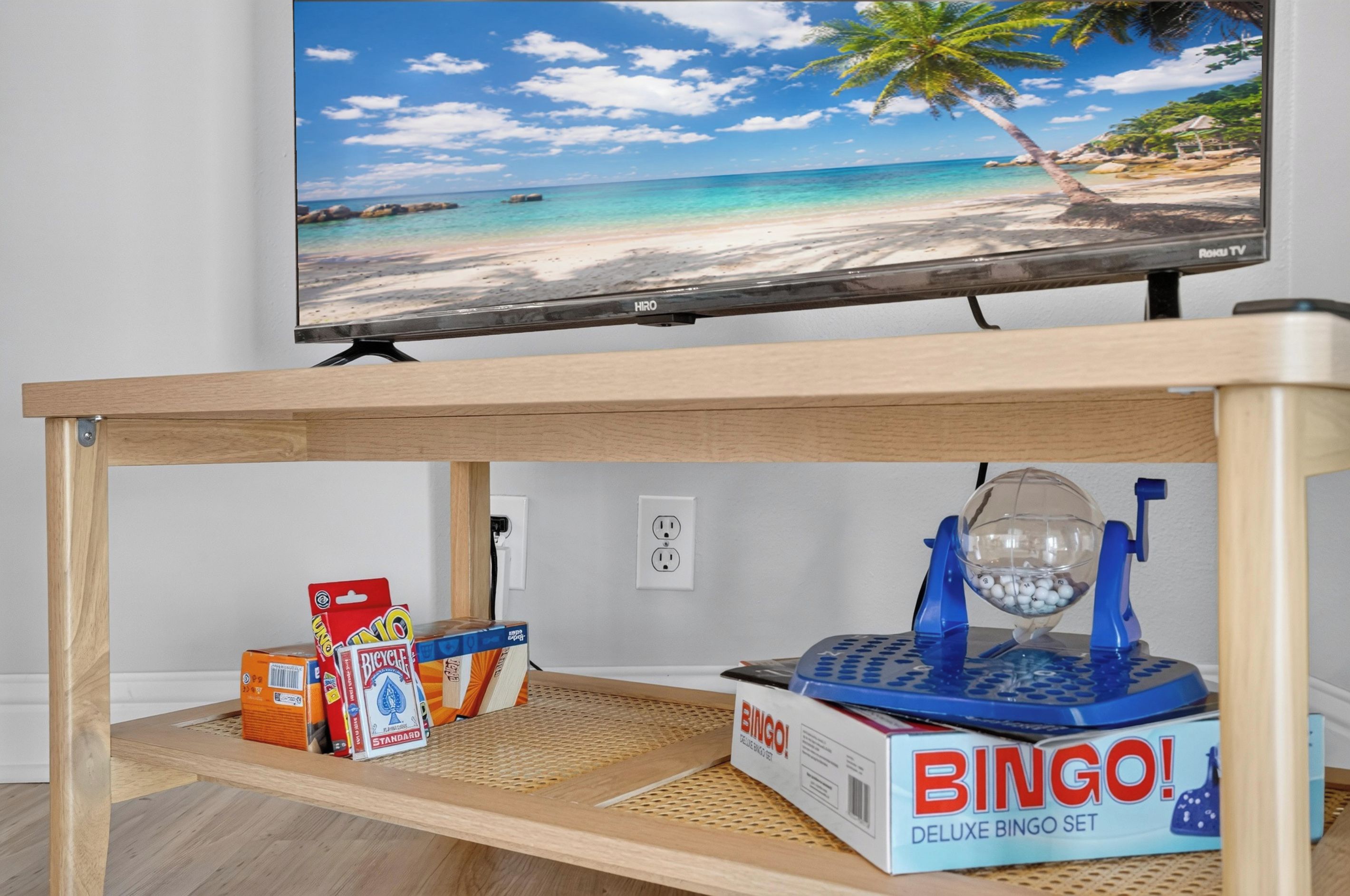 Un téléviseur diffusant une scène de plage, une table en bois sur laquelle est posé un jeu de bingo, et un objet bleu et blanc sur la table.