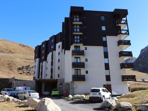 A multi-story building with a stone base and a flat roof, featuring balconies and a parking area with several cars. - Studio cabin near slopes - 4 guests (Tignes)