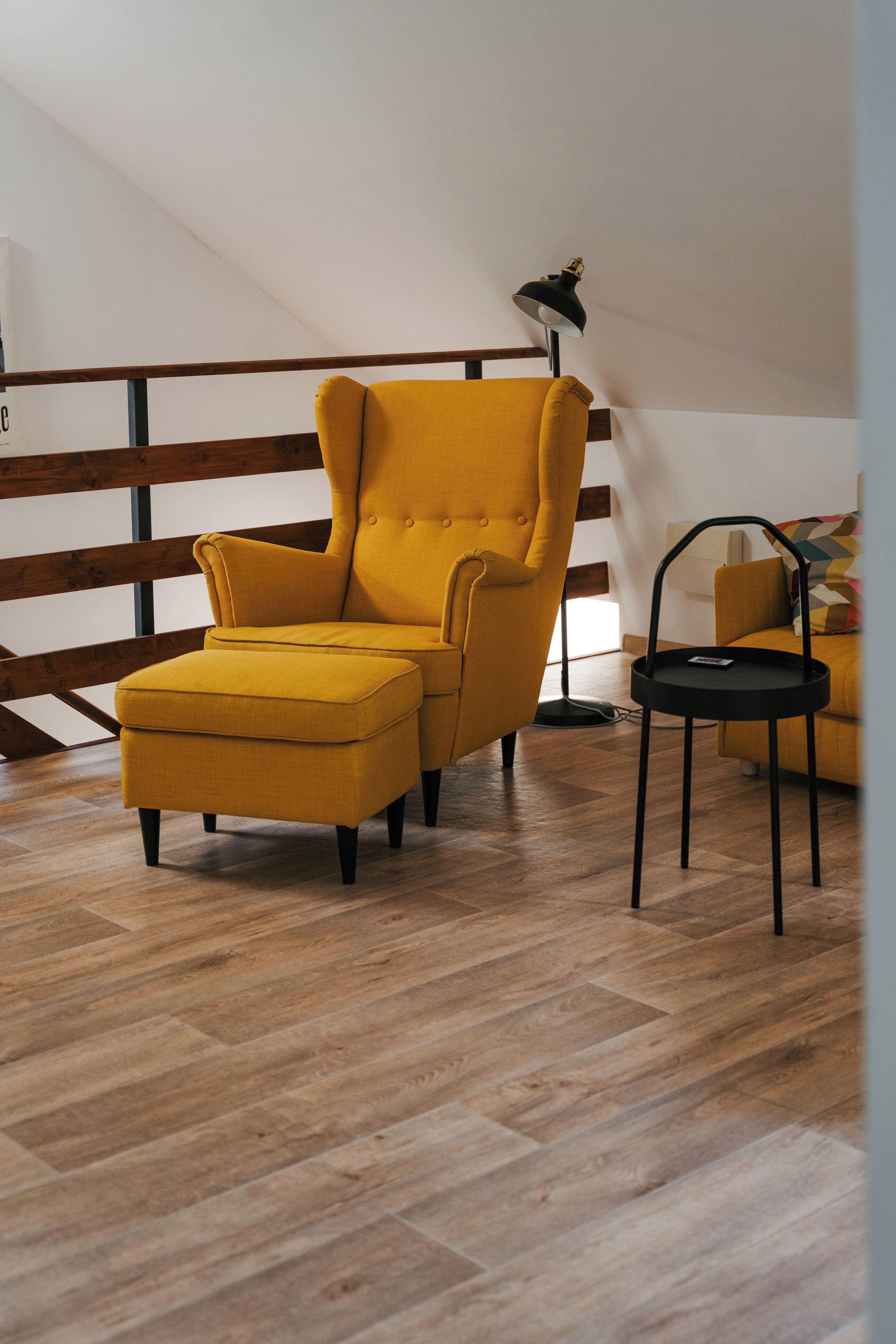 A yellow armchair with a matching ottoman, a small black side table, and a lamp in a room with wooden flooring and a sloped ceiling.