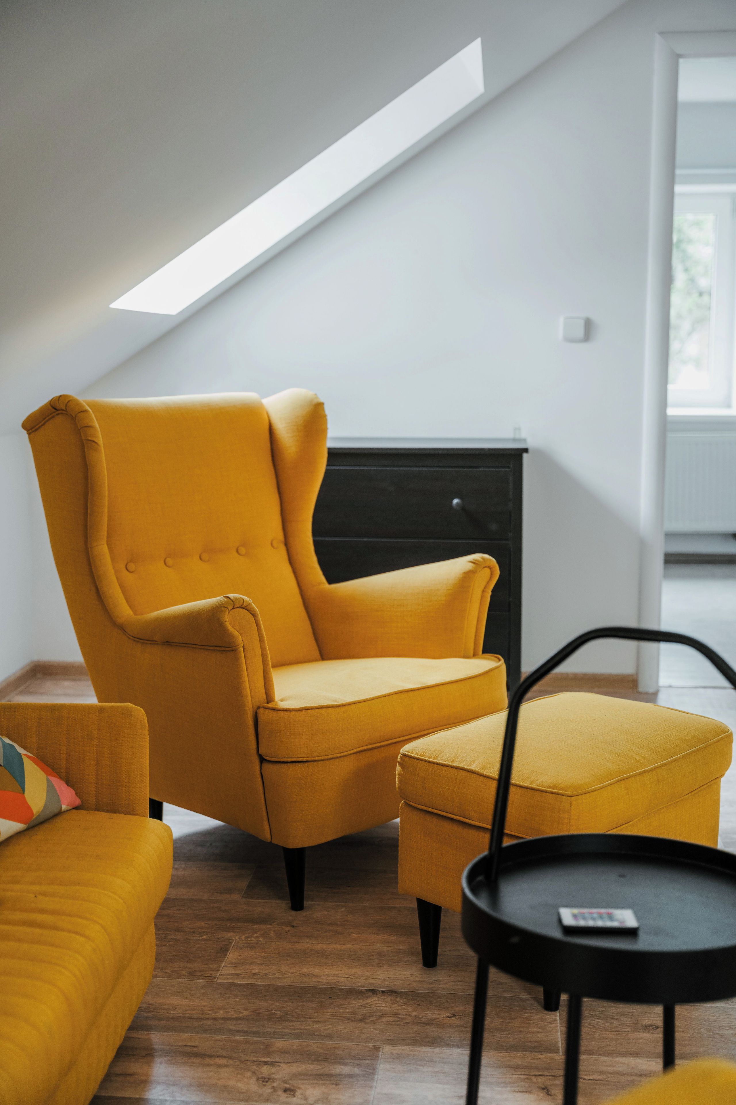 A modern living room with a yellow armchair, a matching ottoman, a black coffee table, and a dark wooden cabinet.