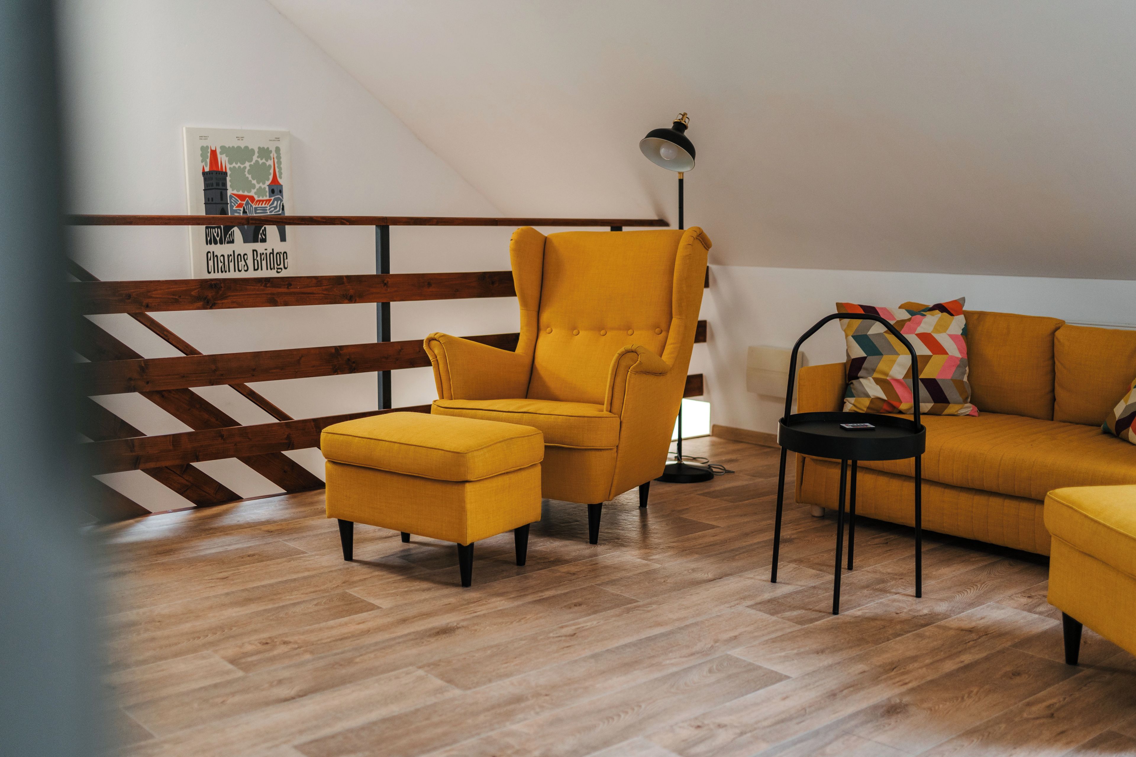 A room with a wooden slatted ceiling, a yellow armchair, a matching ottoman, a black floor lamp, and a sofa with colorful cushions.
