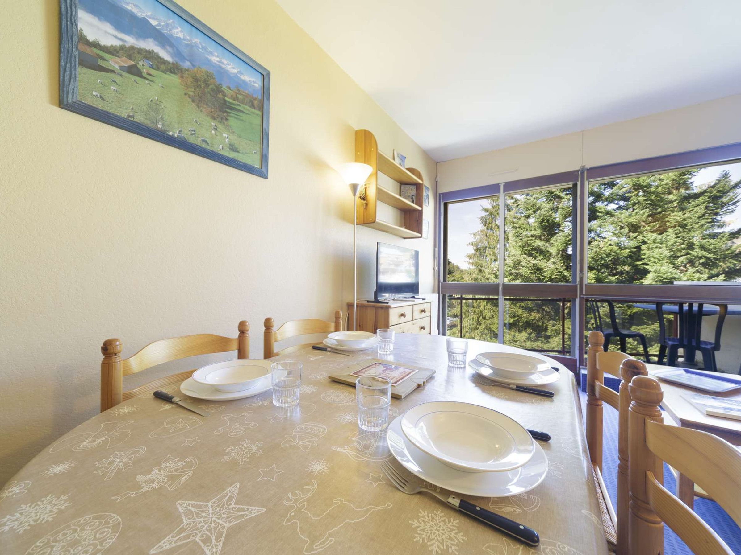 A dining area with a table set for four, chairs, a painting on the wall, and a window with a view of trees.