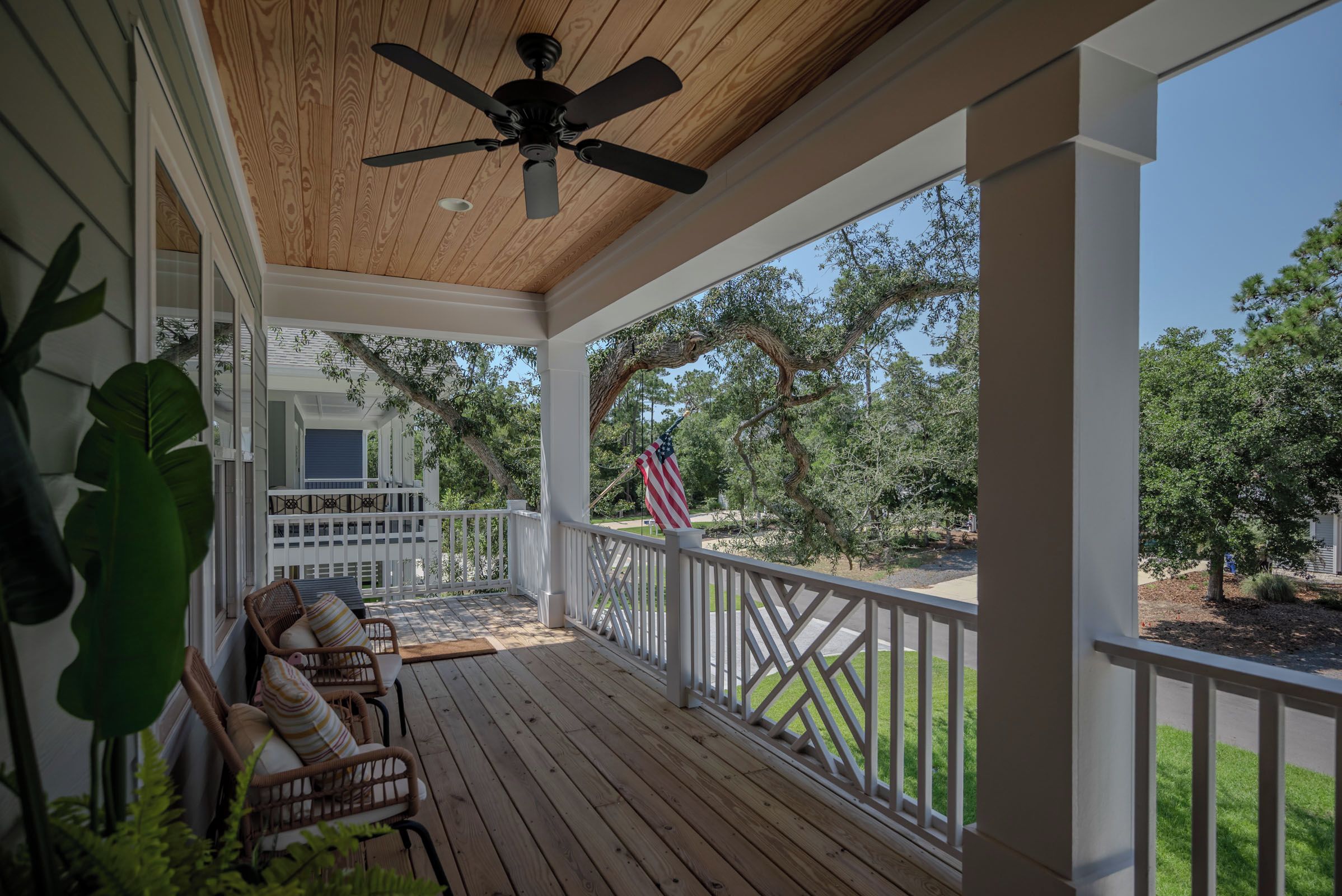 A covered porch with a ceiling fan, wooden deck, white railing, and a view of a residential area with trees and a flag.
