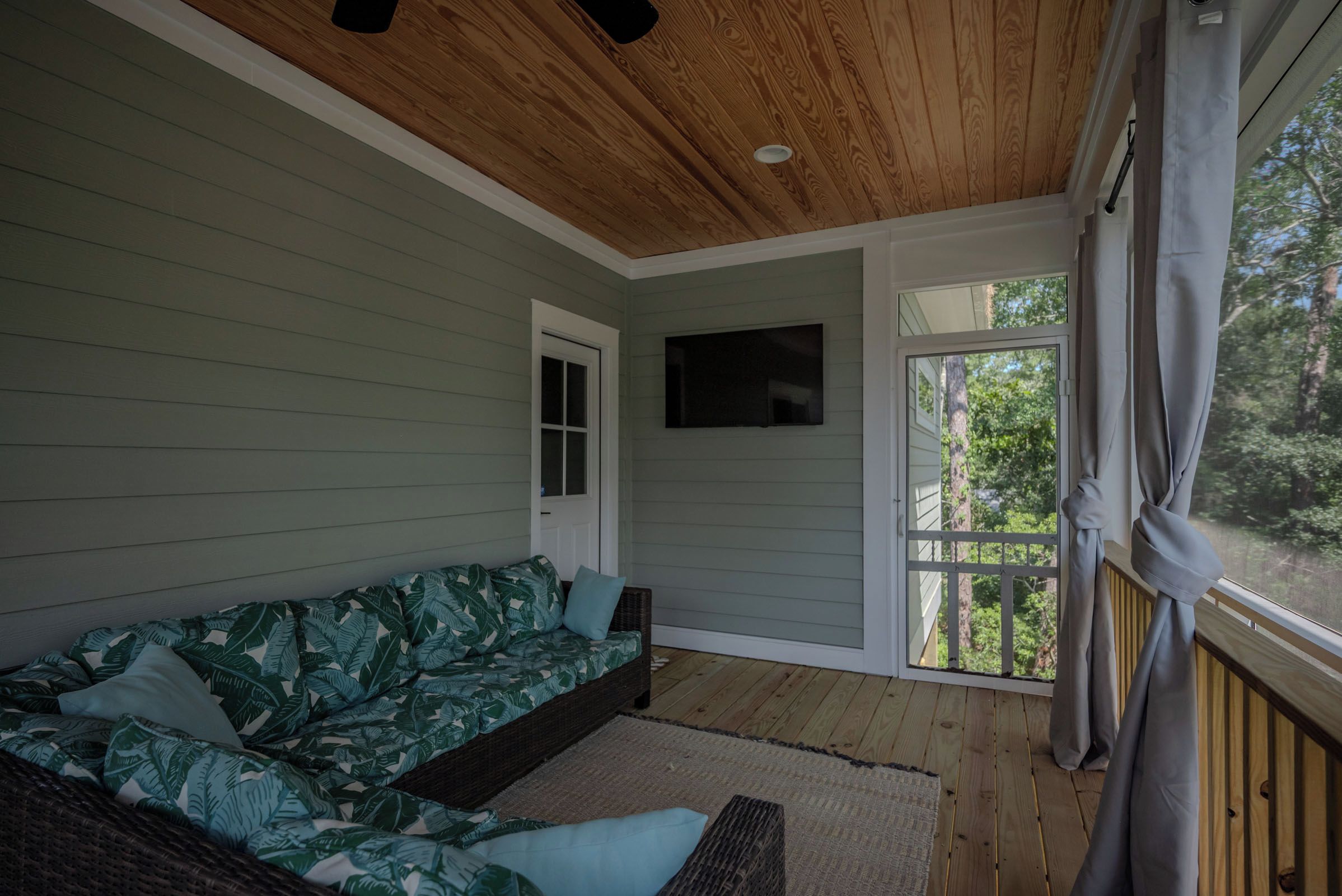 Una habitación con techo de madera, un sofá verde con cojines, un televisor de pantalla plana en la pared y una ventana con vistas a árboles.
