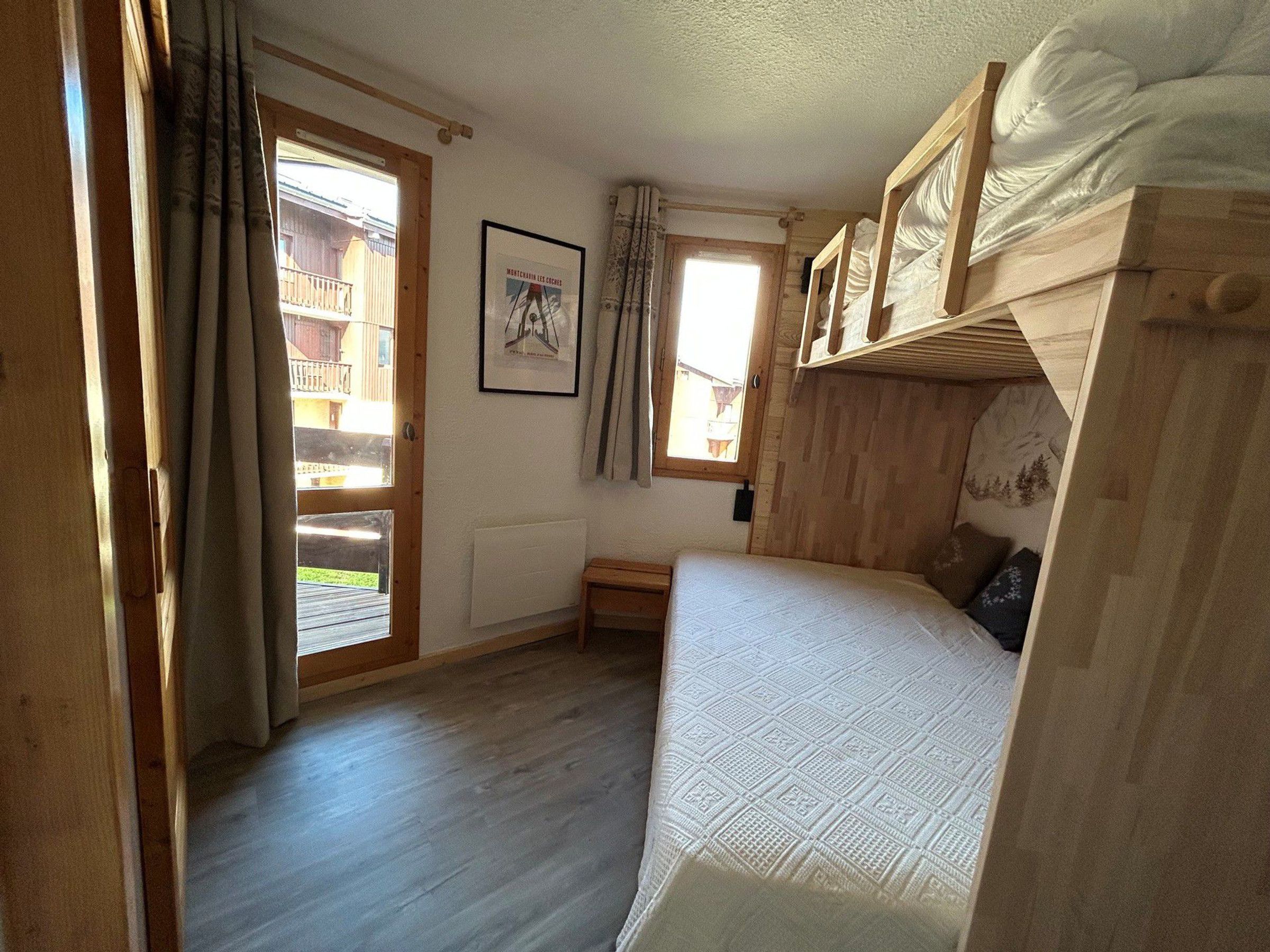 Een kamer met een houten stapelbed, een klein nachtkastje, een ingelijst kunstwerk aan de muur, en een raam met gordijnen.