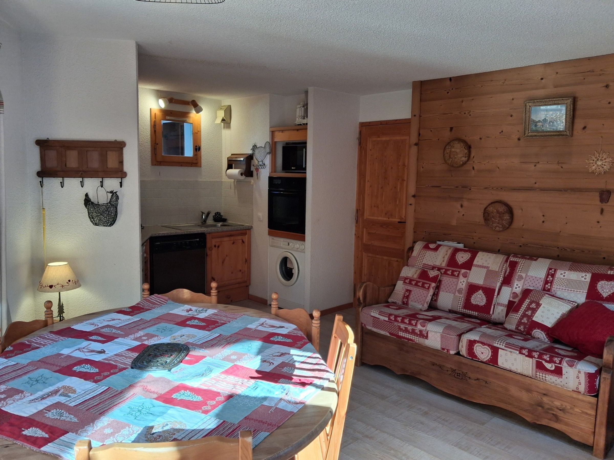 Une cuisine compacte équipée d’un évier, d’une cuisinière et d’un lave-linge. On y trouve une table à manger en bois avec des chaises, ainsi qu’un canapé recouvert d’un tissu à motifs.