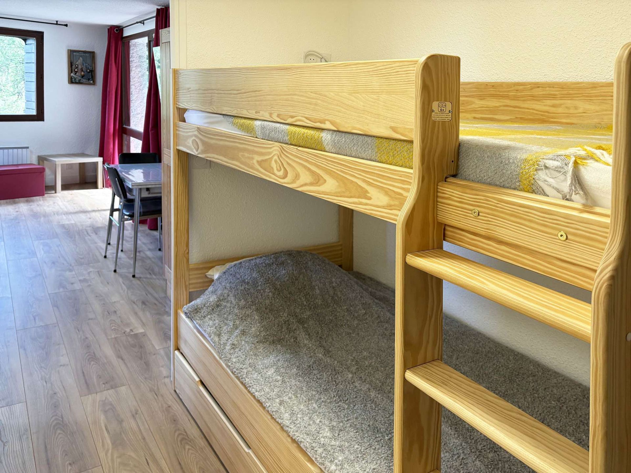 Ein Stockbett mit Holzrahmen und einer grauen Matratze, ein kleiner Tisch und ein Stuhl in einem Zimmer mit Holzboden und einem Fenster mit Vorhängen.