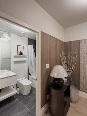 Casa de banho moderna com sanita, lavatório e zona de duche. Há um candeeiro decorativo numa mesa lateral.