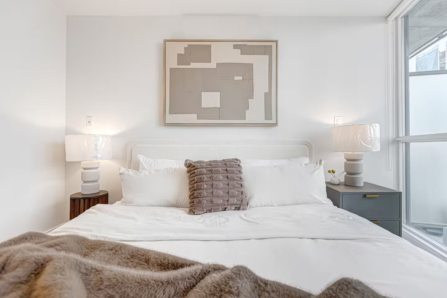A modern bedroom with a large bed, two bedside tables with lamps, a geometric wall art piece, and a view of the outside through a window.