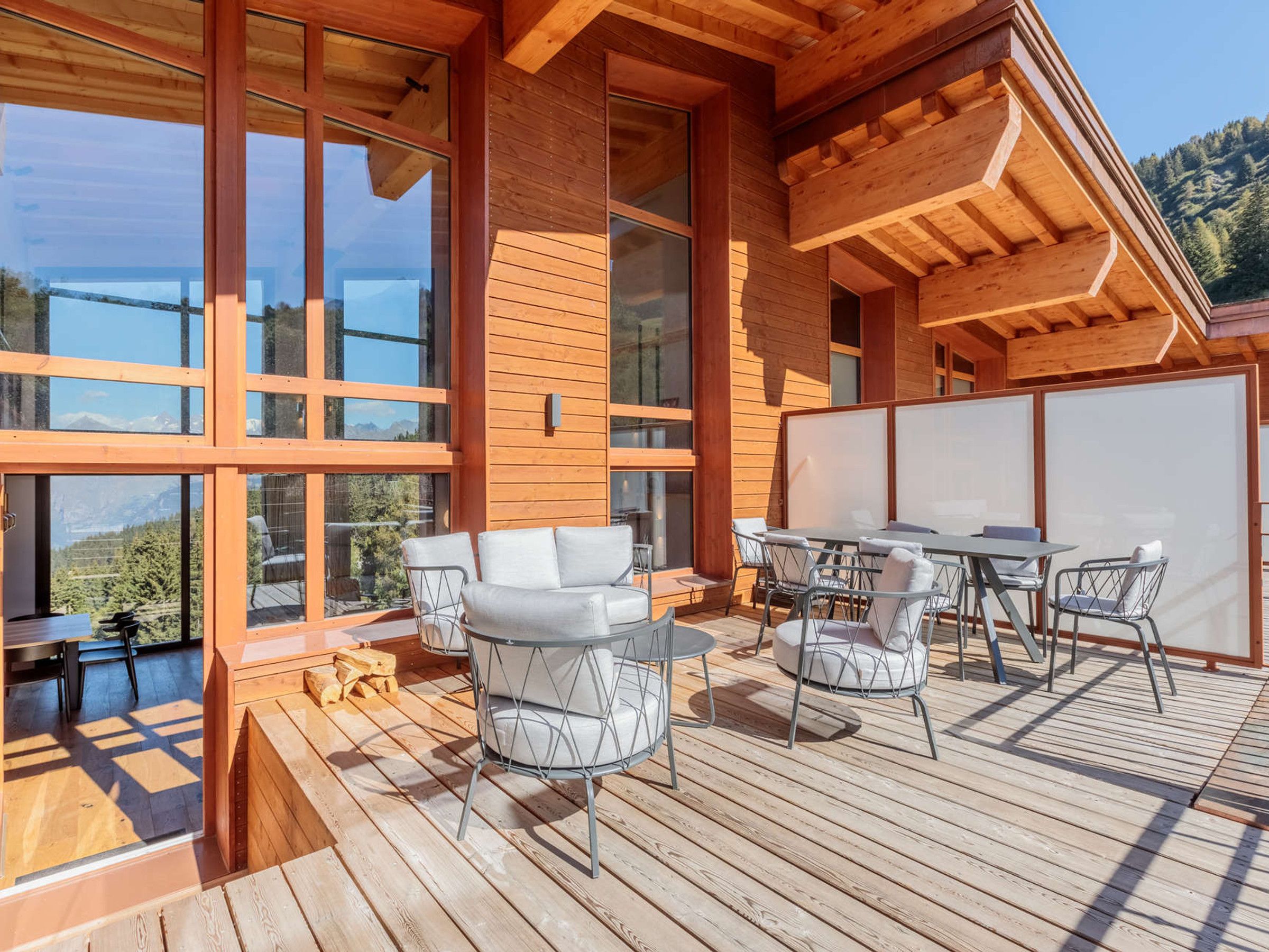 A modern wooden deck with outdoor furniture, large windows, and a view of the mountains.
