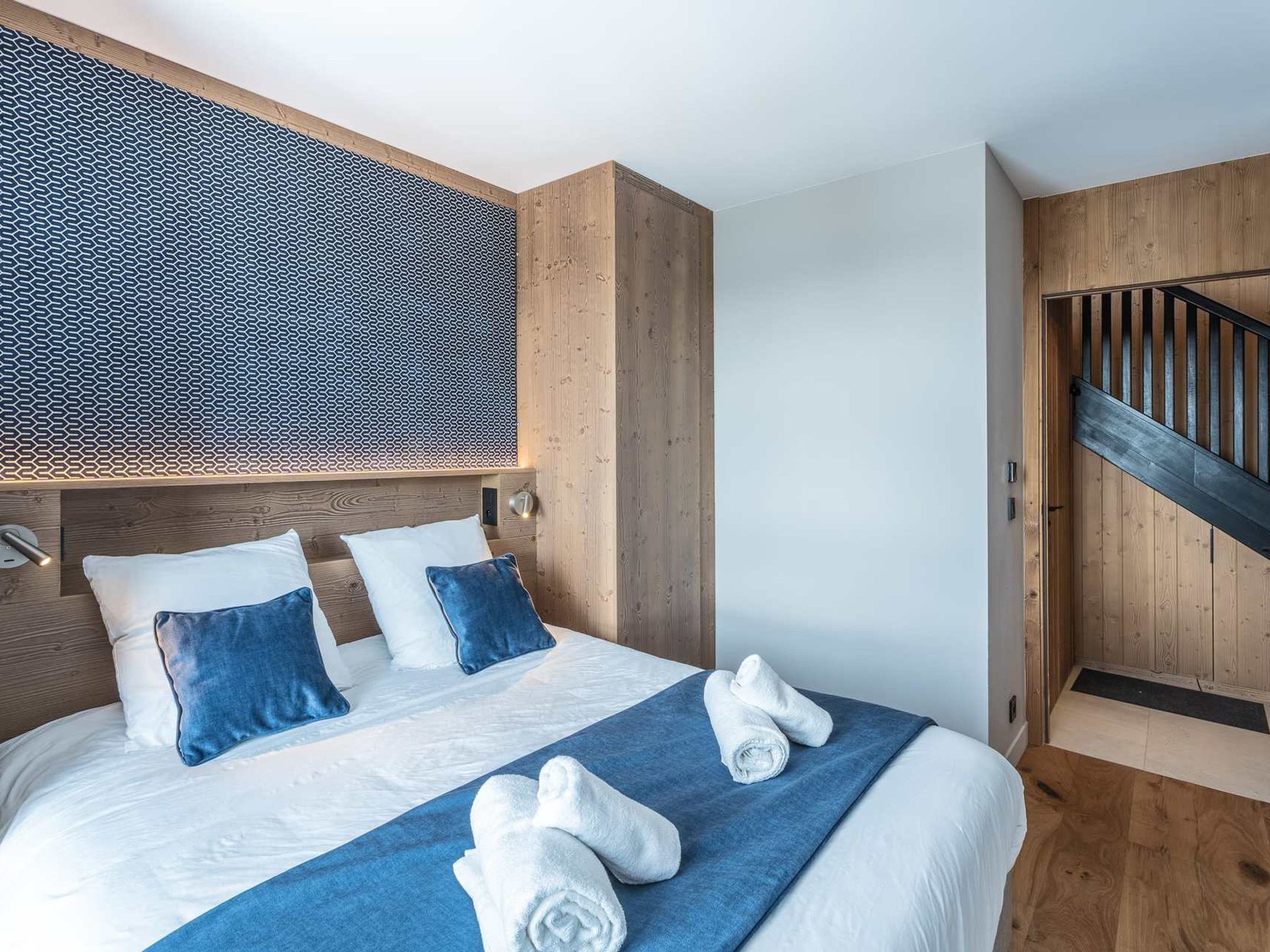 A modern hotel room with a large bed, wooden headboard, and a unique geometric-patterned wall.