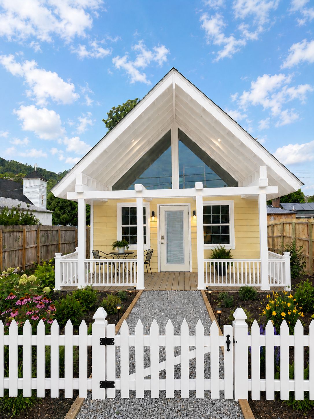 Une maison avec un toit à pignon, une véranda à l’avant avec des balustrades blanches et une clôture en bois peinte en blanc.