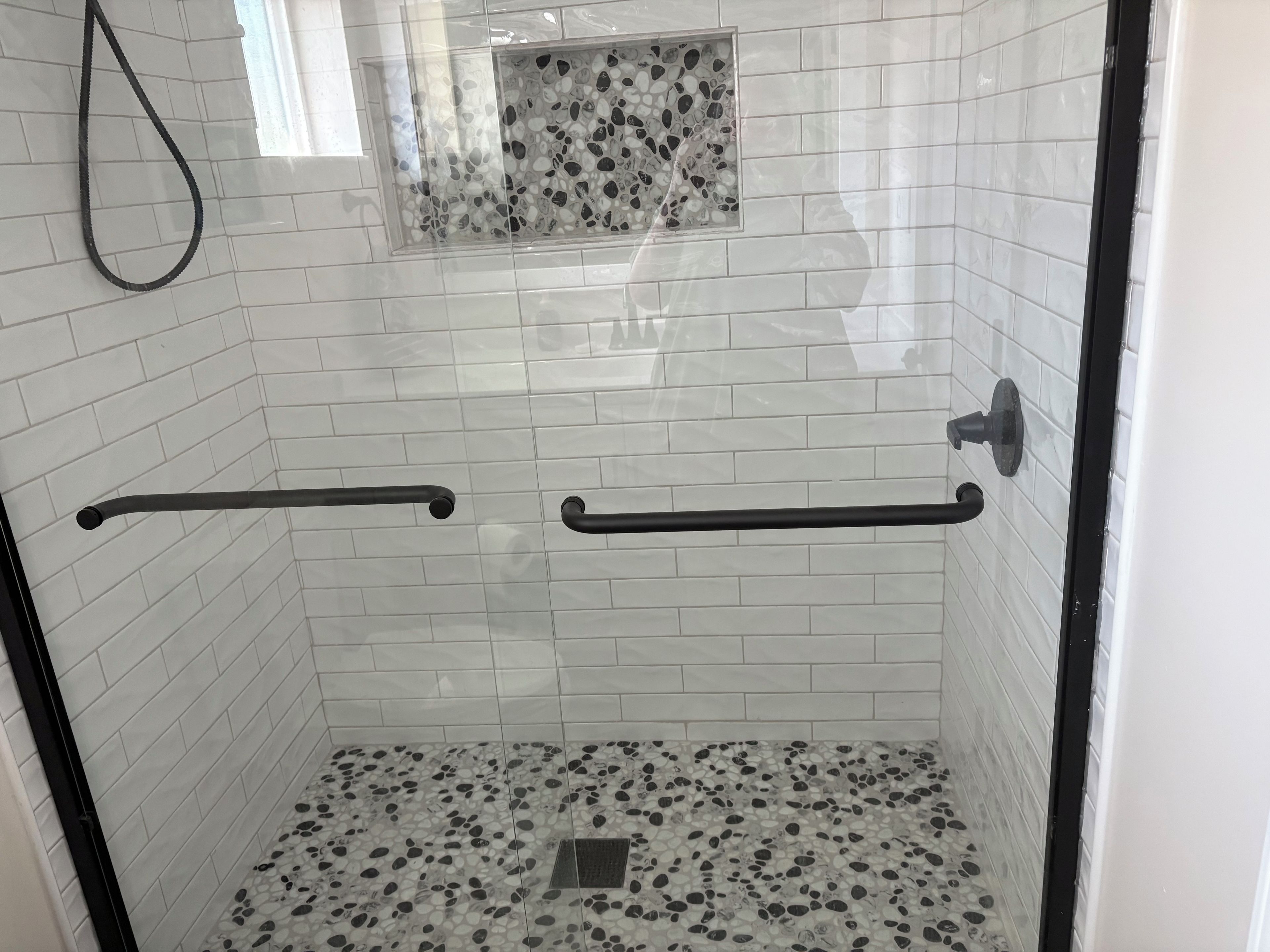 Une douche aux murs recouverts de carreaux blancs, un sol à motifs de galets et une petite fenêtre ornée d’un motif de galets.