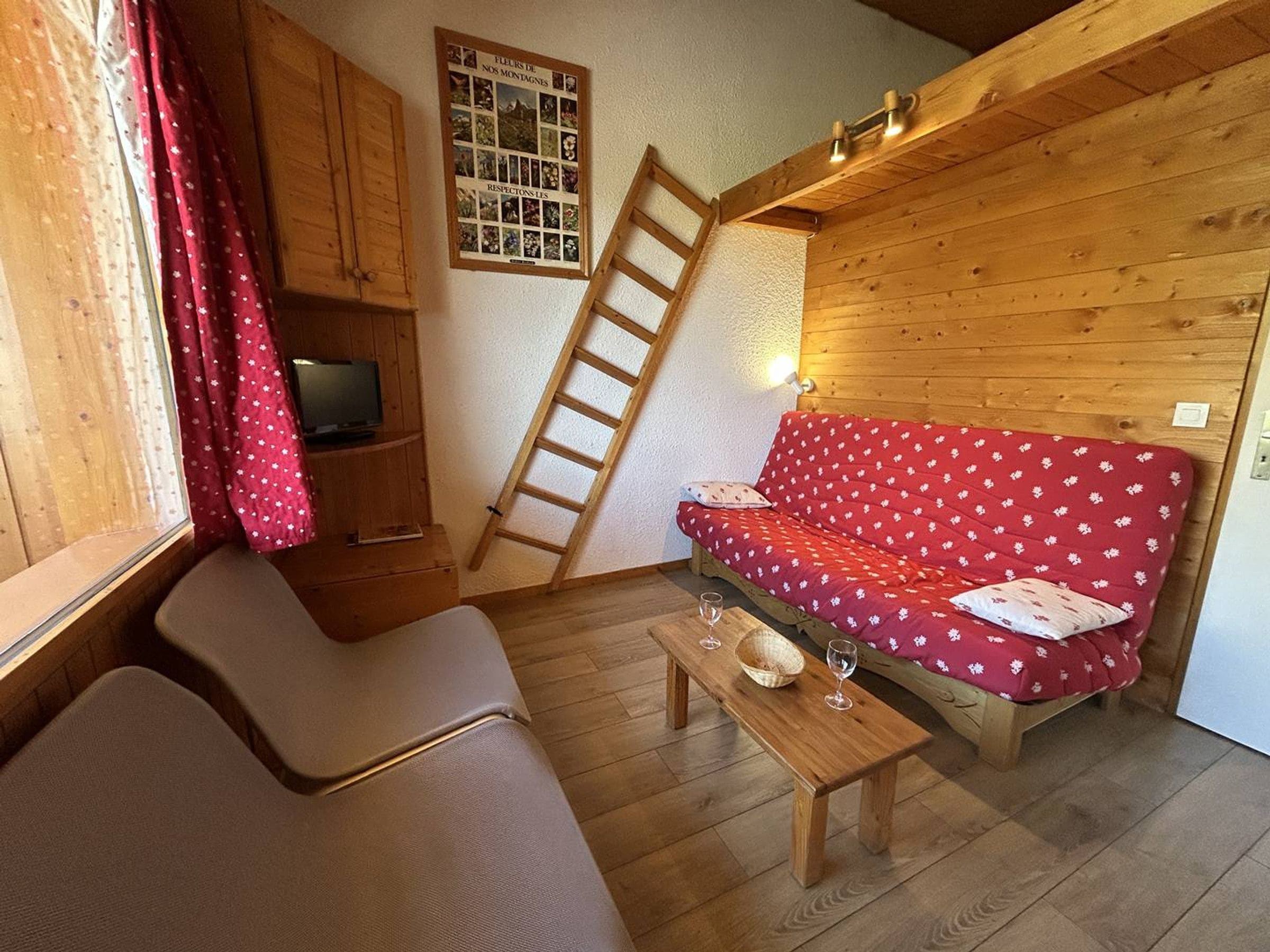 L’intérieur chaleureux d’un chalet avec un canapé, un banc en bois, une échelle et une étagère fixée au mur, sur laquelle se trouve un téléviseur.