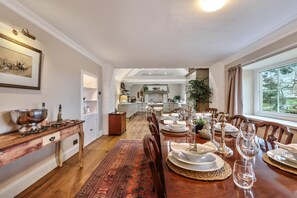Een eetgedeelte met een lange tafel gedekt voor een maaltijd, een keuken op de achtergrond en een groot raam met uitzicht.
