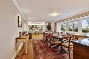 A spacious dining area with a long wooden table and chairs, a kitchen in the background, and large windows offering a view of the outdoors.