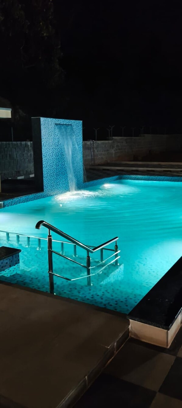 A nighttime swimming pool with a waterfall feature and a metal handrail.