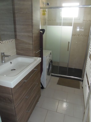 A bathroom with a sink, a washing machine, and a glass shower enclosure. - Vrbo Property (Grandcamp-Maisy)